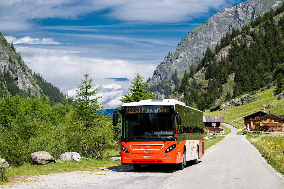 Bus TMR orange circulant sur une route de montagne bordée de chalets