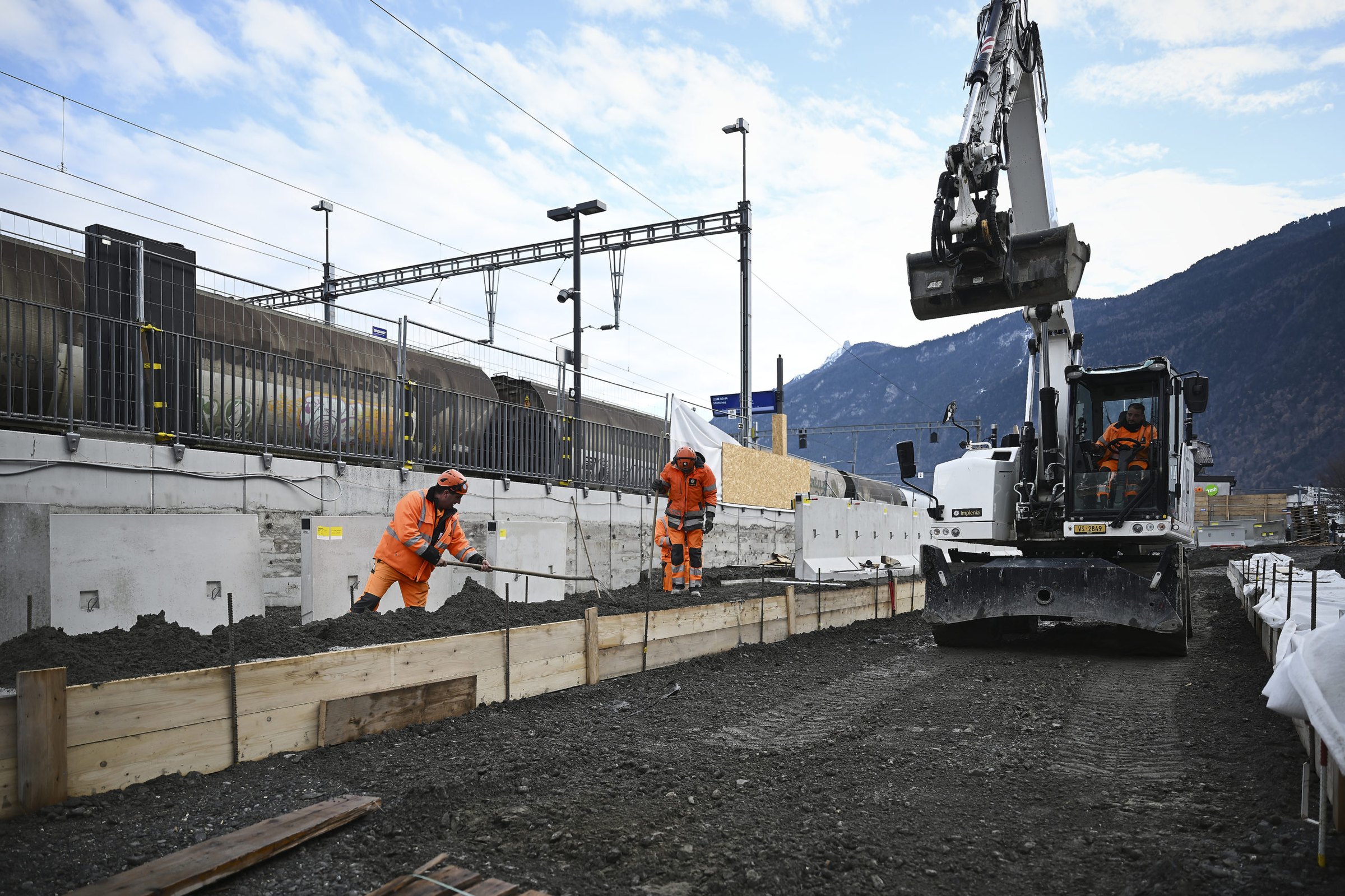 Ouvriers en tenue orange et pelle mécanique sur un chantier de rénovation de gare en Valais